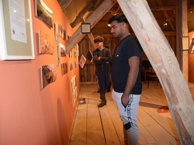 Besuch der Sonderausstellung 'Lehmbau an der Hochschule' im Lehmmuseum Gnevsdorf Ein junger Mann betrachtet die Sonderausstellung 'Lehmbau an der Hochschule' im Lehmmuseum Gnevsdorf. Er steht vor einem gerahmten Foto udn betrachtet es. Im Vordergrund sieht man die Dachkonstruktion aus Holz